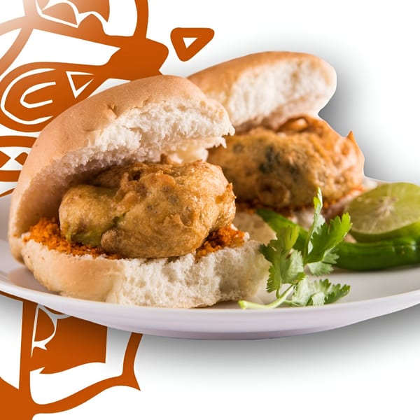 Two vada pavs on a white plate with cilantro, green chili, and lime.